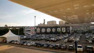 Imagem do estacionamento da Neo Química Arena repleto de veículos. Também é possível ver um muro com pinturas dos escudos do Corinthians